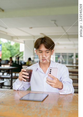 Non binary university student sitting in college campus using phone 128540269