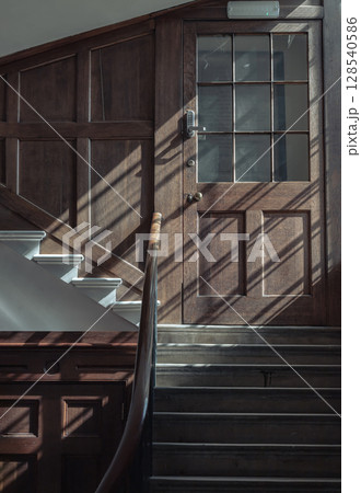 Sunlight streams through the large window, casting light and shadow patterns on surface of wooden door and staircase. Sunlight streams through the large window, casting light and shadow patterns on surface of wooden door and staircase. 128540586