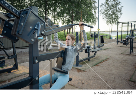 Young woman training on outdoor gym machine in park on a sunny day Young woman training on outdoor gym machine in park on a sunny day 128541394