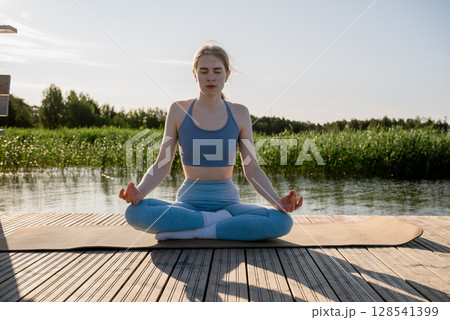 Young woman practicing yoga pose on wooden dock near lake in summer morning light 128541399