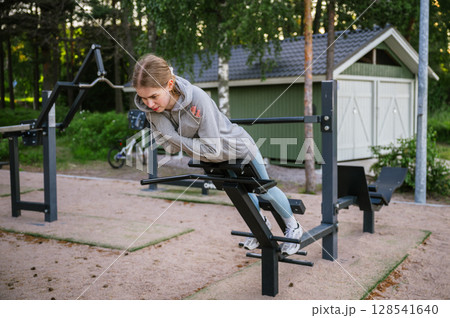 Young woman doing back extension exercise on outdoor fitness bench in summer park 128541640