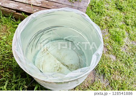 Filling rain barrel lined with transparent plastic liner placed on grass in garden with water collected for further outdoor use. 128541886