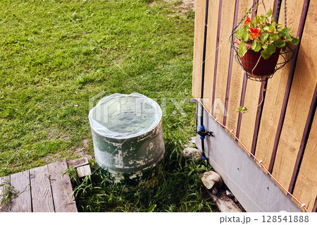 Rain barrel with cistern liner used for garden irrigation placed beside building below roof edge for harvested rain storage 128541888