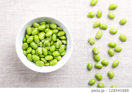 Edamame beans in a white bowl on linen. Shelled young, immature soybeans, Glycine max, of green color, often eaten as snack, steamed or boiled. Popular side dish and appetizer in East-Asian cuisine. Edamame beans in a white bowl on linen. Shelled young, immature soybeans, Glycine max, of green color, often eaten as snack, steamed or boiled. Popular side dish and appetizer in East-Asian cuisine. 128541890
