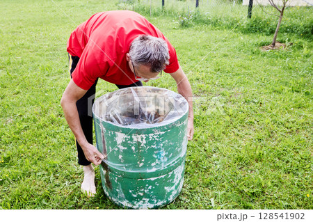 Pulling plastic liner tightly inside rain barrel, gardener ensures liner fits snugly along inner surface of watering barrel in backyard garden. 128541902