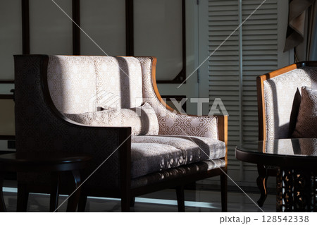 Sunlight shines on the elegant wooden sofa upholstered with ivory velvet fabric and a round coffee table set in a living room. Sunlight shines on the elegant wooden sofa upholstered with ivory velvet fabric and a round coffee table set in a living room. 128542338
