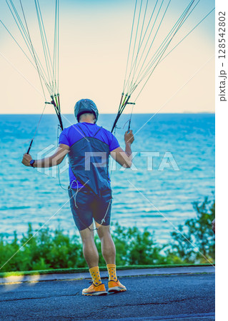 Man preparing to paraglide by sea. Male paraglider holds control lines facing ocean, preparing for takeoff in coastal setting. Concept of paraglider anticipation and sea breeze start 128542802