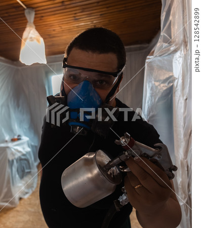 A male painter in protective gear looks at the camera and skillfully operates a spray gun in a room. Vertical, advertising. Surrounded by film, the painter is focused on the accuracy of his painting. A male painter in protective gear looks at the camera and skillfully operates a spray gun in a room. Vertical, advertising. Surrounded by film, the painter is focused on the accuracy of his painting. 128542899