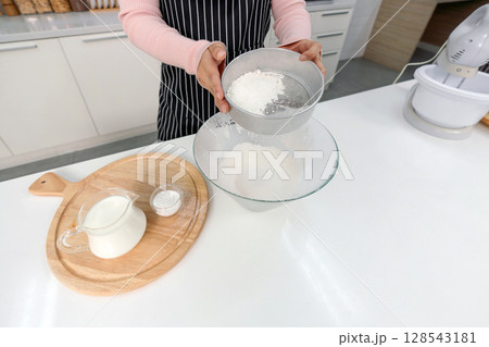 Hand sifting flour in a kitchen with baking ingredient. Baking powder, a jar of milk and electric mixer on the side. 128543181
