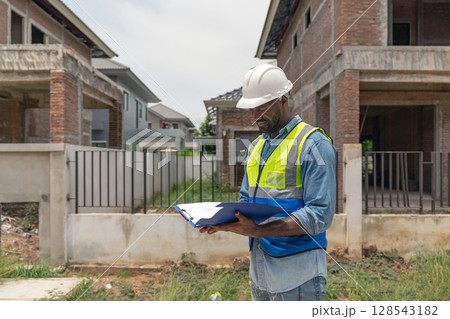 A construction worker in a safety vest and helmet is examining blueprint at a residential building site. Newly built structure surround him, showcasing ongoing construction work. 128543182