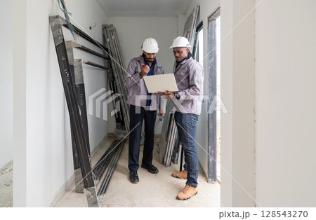 Two construction manager examine plan inside a developing project site. Various construction material surround them, indicating ongoing work. The early morning light create a focused atmosphere. 128543270