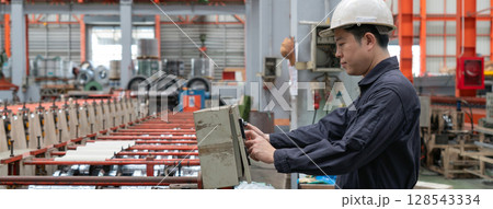 A man in a hardhat and mechanic coverall dust protection suit operates industrial equipment inside a busy factory setting, skillfully managing the machinery. A man in a hardhat and mechanic coverall dust protection suit operates industrial equipment inside a busy factory setting, skillfully managing the machinery. 128543334