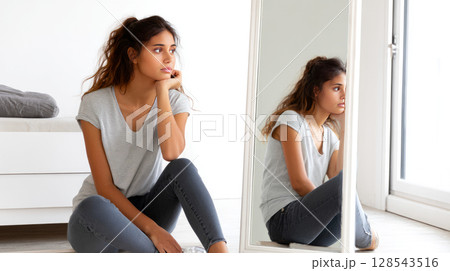 A young girl sitting in front of a mirror, worried about her weight after seeing herself in tight clothes. A young girl sitting in front of a mirror, worried about her weight after seeing herself in tight clothes. 128543516