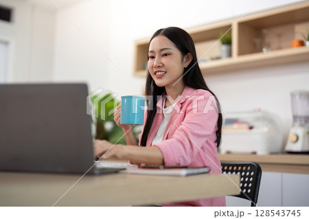 Working from Home: Young Woman Typing on Laptop with Coffee in Hand Working from Home: Young Woman Typing on Laptop with Coffee in Hand 128543745