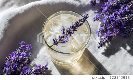 A refreshing drink with ice cubes and a sprig of lavender on a soft white fabric. The scene is bright and inviting, perfect for summer relaxation. 128543938