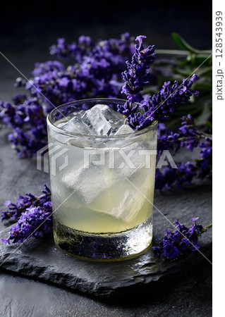 A refreshing cocktail with ice cubes in a glass, surrounded by fresh lavender flowers on a dark slate surface. The drink has a light yellow color. 128543939