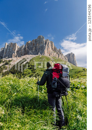Man hiker with trekking poles, backpack and solar panel stands in green meadow with rocky mountains Man hiker with trekking poles, backpack and solar panel stands in green meadow with rocky mountains 128544296