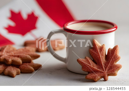 A white mug filled with coffee sits beside maple leaf-shaped cookies. A Canadian flag is in the background, symbolizing Canada Day celebrations on July 1st. A white mug filled with coffee sits beside maple leaf-shaped cookies. A Canadian flag is in the background, symbolizing Canada Day celebrations on July 1st. 128544455