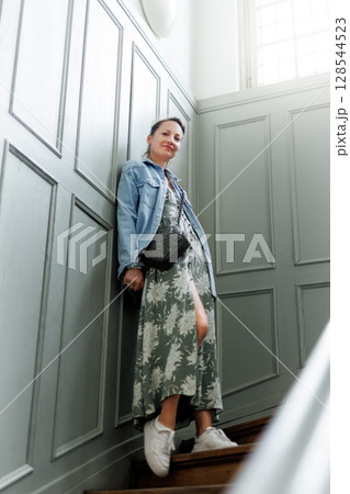 Stylish woman in floral dress and denim jacket stands on wooden staircase beside vintage paneled wall. Soft light from large window highlights her calm pose and relaxed, confident expression Stylish woman in floral dress and denim jacket stands on wooden staircase beside vintage paneled wall. Soft light from large window highlights her calm pose and relaxed, confident expression 128544523
