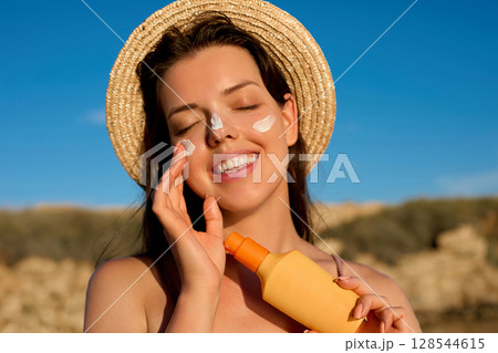 Beautiful Young woman with sun cream on face. Girl holding sunscreen bottle on the beach. Female in hat applying moisturizing lotion on skin.Skin care. Sun protection. Suntan. Beautiful Young woman with sun cream on face. Girl holding sunscreen bottle on the beach. Female in hat applying moisturizing lotion on skin.Skin care. Sun protection. Suntan. 128544615