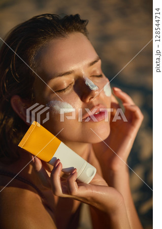 Smiling happy woman applying sunscreen to her face on the beach at sunset.  Cream, moisturizer and facial woman in skincare, beauty or cosmetics. 128544714