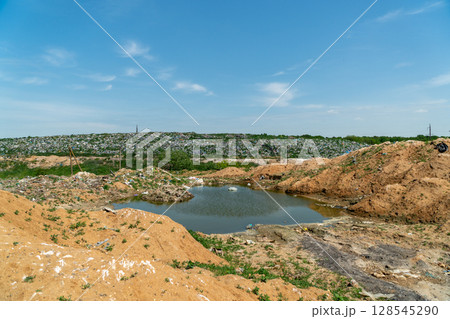 A wasteland of garbage rises in the background with a small pond and patches of greenery under a bright sky A wasteland of garbage rises in the background with a small pond and patches of greenery under a bright sky 128545290