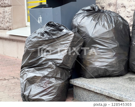 Piles of black trash bags are leaning against a stone surface next to recycling bins on a busy city sidewalk in the afternoon sunlight Piles of black trash bags are leaning against a stone surface next to recycling bins on a busy city sidewalk in the afternoon sunlight 128545303