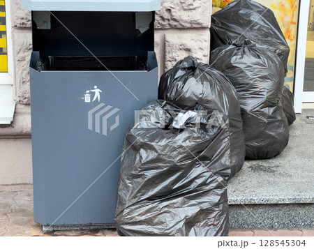 Black trash bags pile up beside an overflowing public garbage bin in a city setting, highlighting waste management issues in urban areas Black trash bags pile up beside an overflowing public garbage bin in a city setting, highlighting waste management issues in urban areas 128545304