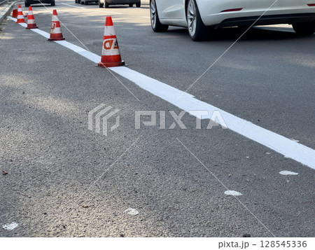 Traffic cones surround freshly painted white lines on an urban street during daytime as vehicles pass nearby 128545336