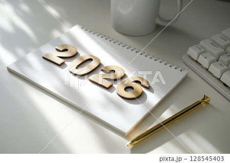 Golden numbers 2026 on notebook, golden pen, white mug and keyboard part, bathed in soft sunlight. Planning, goals, new beginnings. Ideal for business, education 128545403