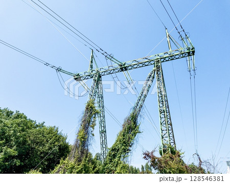 Electric power transmission towers with high-voltage lines against clear blue sky, metal lattice structures for energy distribution, industrial infrastructure in outdoor urban environment Electric power transmission towers with high-voltage lines against clear blue sky, metal lattice structures for energy distribution, industrial infrastructure in outdoor urban environment 128546381