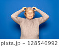 Caucasian woman in her 70s wearing a dark beige sweater scratching her head worriedly trying to remember something important, isolated over blue background. 128546992