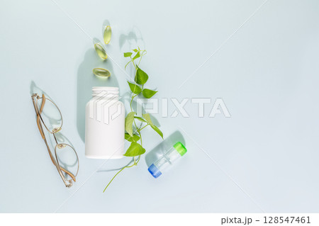 Mockup bottle with natural pills or vitamins for eye health on blue background. White bottle with healthy supplements in gel capsules with glasses and contact lenses. Good vision concept. Copy space 128547461
