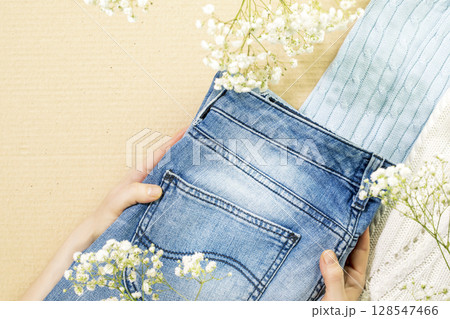 Second hand concept. Sustainable life style with female hands holding jeans near sweaters on cardboard background. Circular fashion, eco friendly sustainable shopping, thrifty shop concept. Copy space 128547466