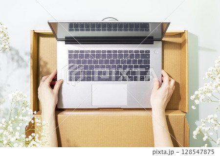 Buying and delivering new laptop. Romantic overhead shot of womans hands holding laptop in cardboard box on blue background with white spring flowers and shadows. Flat lay 128547875