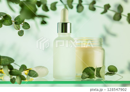 Glass dropper bottle with face serum or oil on glass shelf with candle on blue background with eucalyptus branches and shadows. Natural cosmetics based on fermented products. Front view, soft focus 128547902