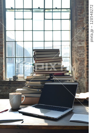 A stack of books and a laptop on a desk with a view of the outside 128548272