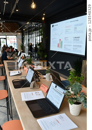 A group of people are sitting at a long table with laptops open in front of them 128548329