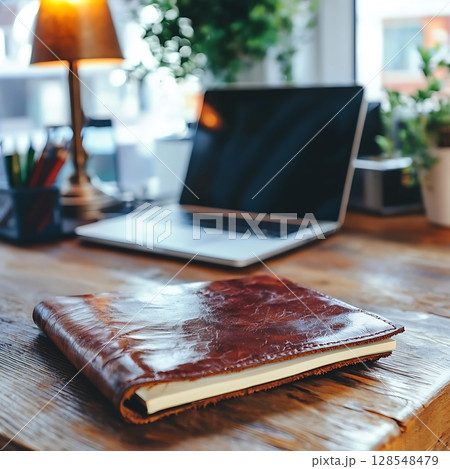 A leather bound notebook sits on a wooden desk next to a laptop 128548479