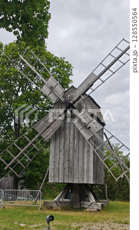 Old wooden windmill with large blades standing on grass under a cloudy sky surrounded by trees 128550564