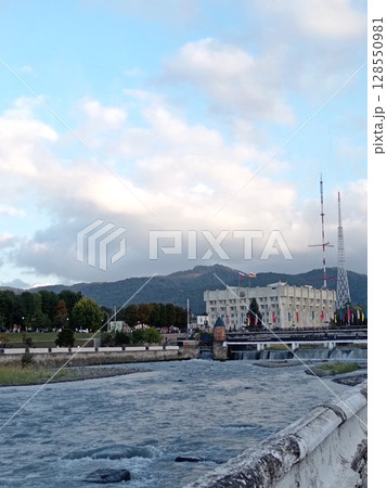 Scenic urban landscape along Terek River: Vladikavkaz City Hall standing proudly on the embankment, Caucasus mountains in background 128550981