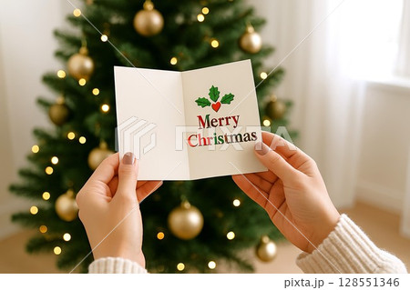 Christmas card in front of decorated tree with festive holiday greeting in women's hands. concept of christmas celebration, holiday spirit, festive greeting card 128551346