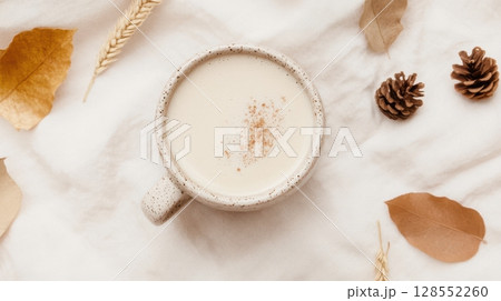 Cozy autumn flatlay with beige chai latte in ceramic mug surrounded by pinecones, wheat, and dried leaves on soft textile with warm lighting 128552260