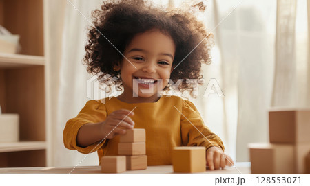 Smiling Black toddler girl plays with wooden blocks at a low table in a cozy room filled with sunlight and natural textures. 128553071