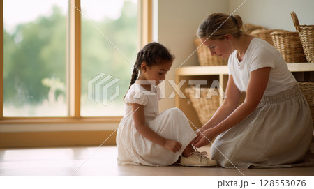 Tender moment of a woman helping a child with shoes in a serene, sunlit room with floor mats and open shelves. Tender moment of a woman helping a child with shoes in a serene, sunlit room with floor mats and open shelves. 128553076