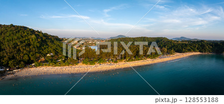 Aerial view of Naiharn Beach in Phuket, Thailand, with turquoise waters, lush green hills, scattered buildings, and a clear blue sky in the background. 128553188