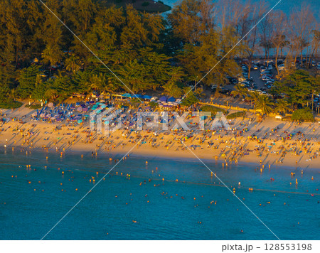 Aerial view of Naiharn Beach in Phuket, Thailand, showing a busy shoreline with umbrellas, swimmers, and a lush green tree line in warm golden light. Aerial view of Naiharn Beach in Phuket, Thailand, showing a busy shoreline with umbrellas, swimmers, and a lush green tree line in warm golden light. 128553198