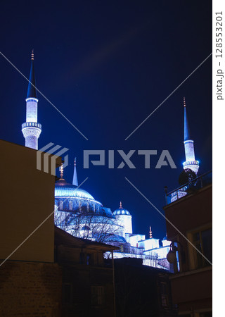 The Blue Mosque in Istanbul, Turkey, glows at night with its domes and minarets framed by nearby buildings and bare tree branches under a deep blue sky. The Blue Mosque in Istanbul, Turkey, glows at night with its domes and minarets framed by nearby buildings and bare tree branches under a deep blue sky. 128553201