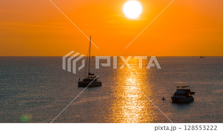 A vibrant sunset at Naiharn Beach in Phuket, Thailand, with orange and yellow hues reflecting on calm water. A sailboat and motorboat are anchored nearby. 128553222
