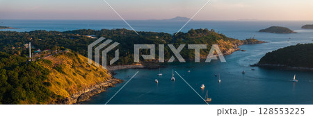 Aerial view of Naiharn Beach in Phuket, Thailand, featuring a wind turbine on a hill, sailboats in the bay, and distant islands under a golden sunset. 128553225